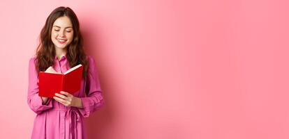 Attractive modern woman writing in her diary, making notes in notebook or planner, looking at her schedule, standing against pink background photo