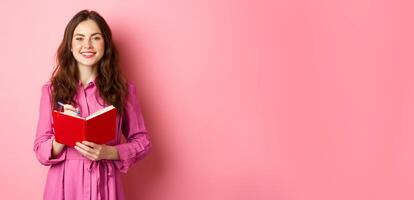 Cheerful girl smiling and writing down notes, holding her planner, plan schedule or grocery list, standing with diary against pink background photo