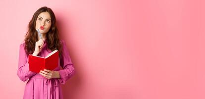 Beautiful young woman look thoughtful, writing in notebook, holding planner or diary, plan her schedule, standing against pink background photo