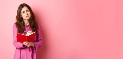 Sad young woman writing her feelings in diary, looking upset and gloomy, feeling heartbroken, sharing memories in planner, standing against pink background photo