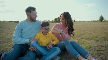 Cute Caucasian happy family portrait joyful man male dad smiling woman female mom little boy child kid son sitting together hug embrace looking camera outside nature field farmland park picnic nature video