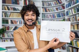 Young arab tutor in headset with microphone holding drawing board with lesson 2 inscription while smiling at camera. Positive man pointing with pen to tablet on background of bookshelves. photo