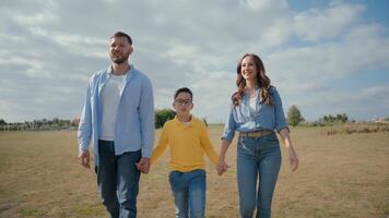 Young loving Caucasian parents female smiling woman mother male man father holding hands happy little boy child adopted kid son talking speak walking outside park field. Family relationship travelling video