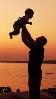 Smiling father throws his little baby boy up in the air. Dad and son having family time outdoors in the nature. River in sunset at backdrop. Vertical video