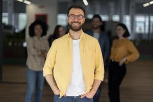 Confident businessman smiling with hands in pockets in office with team in background photo