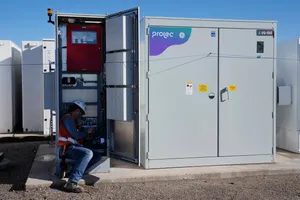 A worker in a hardhat sits on the ground in the doorway of a large, boxy structure somewhat like a storage locker.