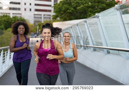 Happy young curvy women jogging together on city bridge. Healthy girls friends running on the city street to lose weight. Group of multiethnic oversize women running with building in the background.