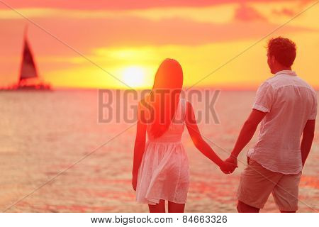 Honeymoon couple romantic in love holding hands at beach sunset. Newlywed happy young couple enjoying ocean sunset during travel holidays vacation getaway. Interracial couple.