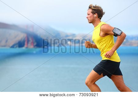 Athlete running man - male runner in San Francisco listening to music on smartphone. Sporty fit young man jogging by San Francisco Bay and Golden Gate Bridge. Jogger training with smart phone armband,