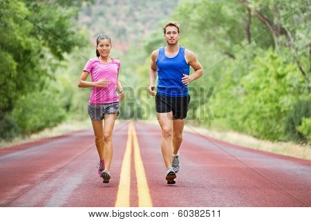 Fitness sport couple running jogging outside on road beautiful nature landscape. Runners training together for marathon run. Asian female sports woman and fit male fitness man in full body length.