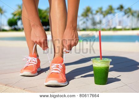 Running woman runner with green vegetable smoothie.  Fitness and healthy lifestyle concept with female model tying running shoe laces.