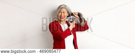 Scared Asian Woman Cutting Hair With Scissors And Feeling Anxious, Close Eyes Nervously, Standing In