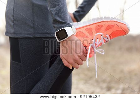 Running stretching - runner wearing smartwatch. Closeup of running shoes, woman stretching leg as warm-up before run with sport activity tracker watch at wrist to monitor the heart rate during cardio.