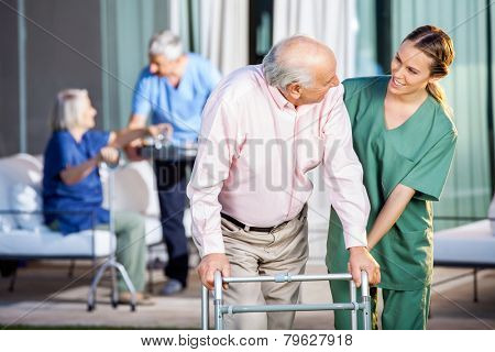 Happy female caretaker assisting senior man in using Zimmer frame at nursing home yard