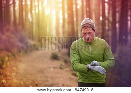 Trail running runner looking at heart rate monitor watch running in forest wearing warm jacket sportswear, hat and gloves. Male jogger running training in woods.