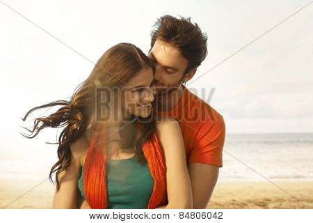 Attractive young casual caucasian brunette woman getting a kiss from boyfriend at beach. Romance, couple, smiling, embracing. Lens flare, sand and copyspace.