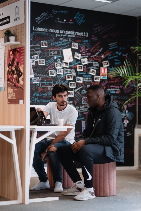 Two young men sit on chairs at a desk looking at a computer screen in front of them. Behind them is a wall covered in brainstorming notes and photos.