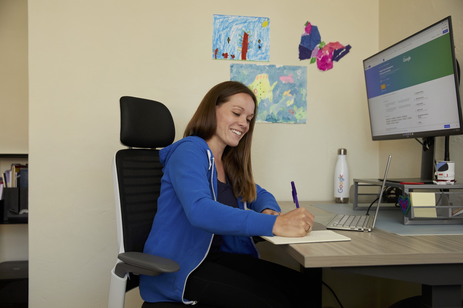 A woman smiles as she makes some notes at her desk, children’s drawings visible on the wall behind her.