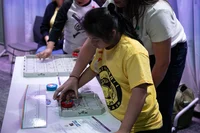 A teacher helps a female middle school student move a vex robot with her hands at a table