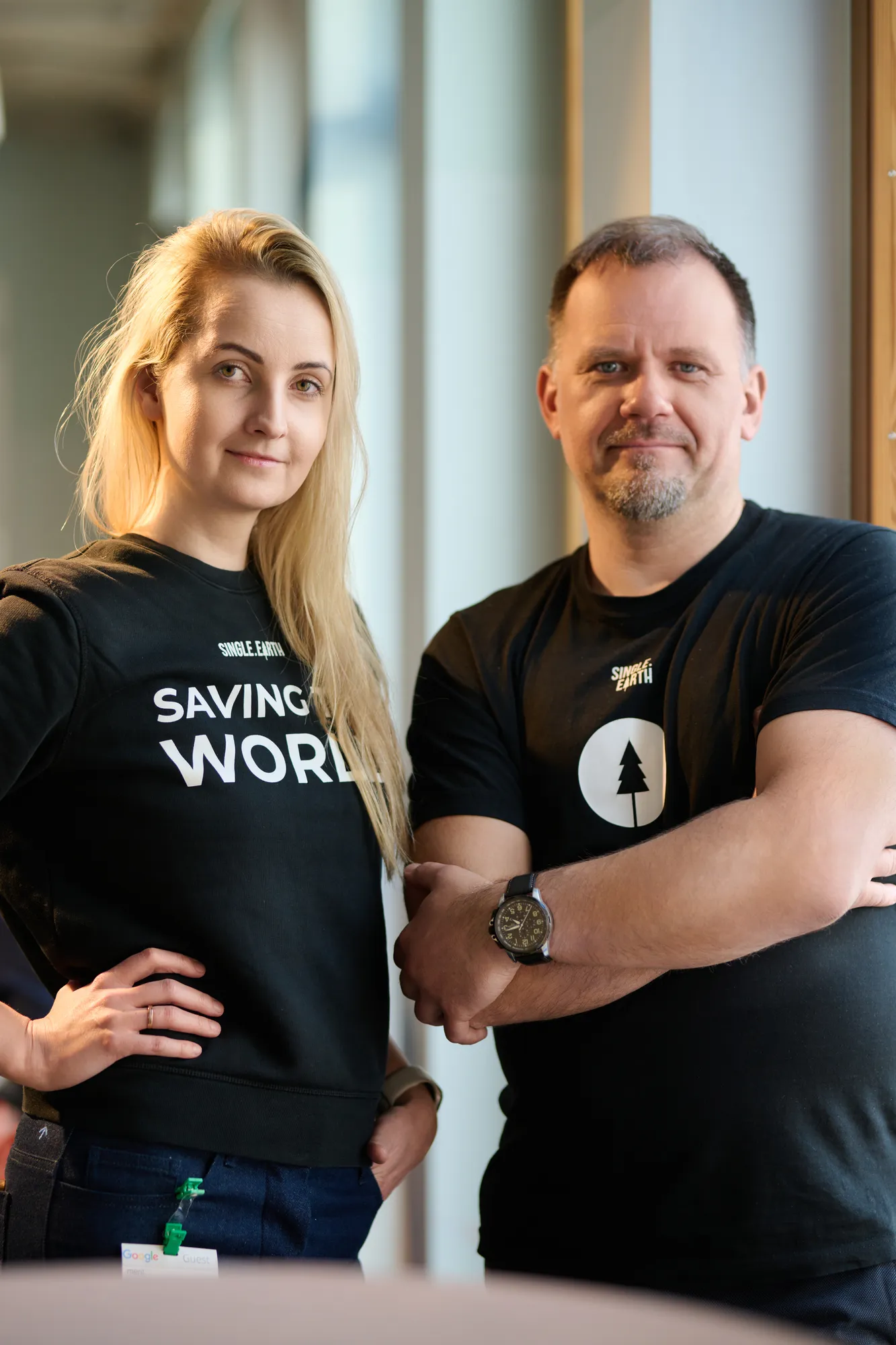 A woman with blonde hair and a man with brown hair wear black t-shirts and stand looking at the camera.