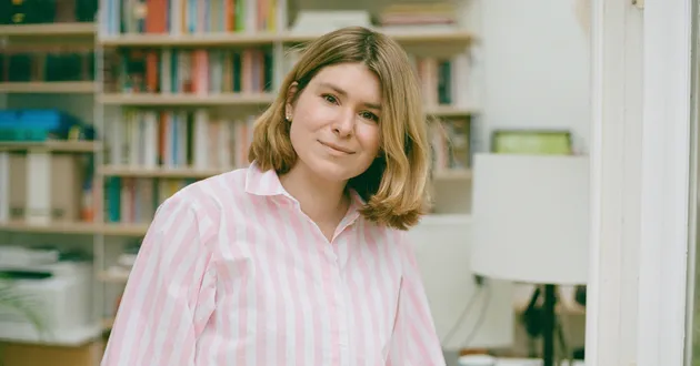 A woman wearing a pink and white striped shirt and with shoulder-length blonde hair looks at the camera, standing in front of a bookshelf.