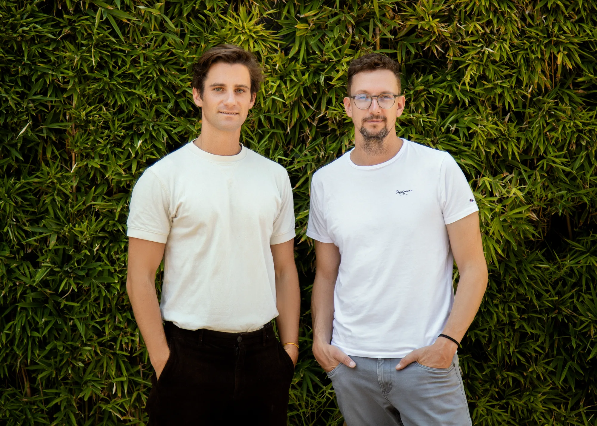 Two men wearing white t-shirts stand looking at the camera in front of a living wall.