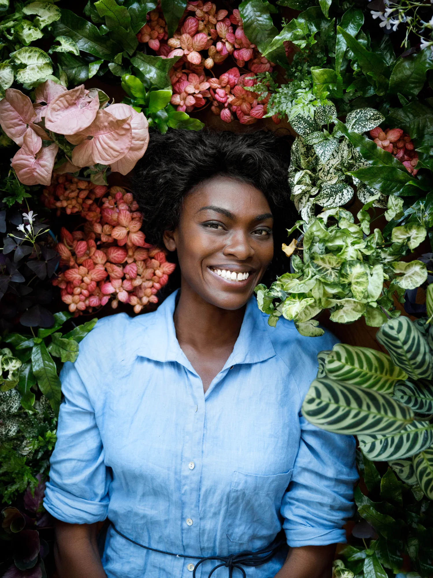 Photo showing Deborah Choir smiling looking at the camera against a wall of flowers and plants.