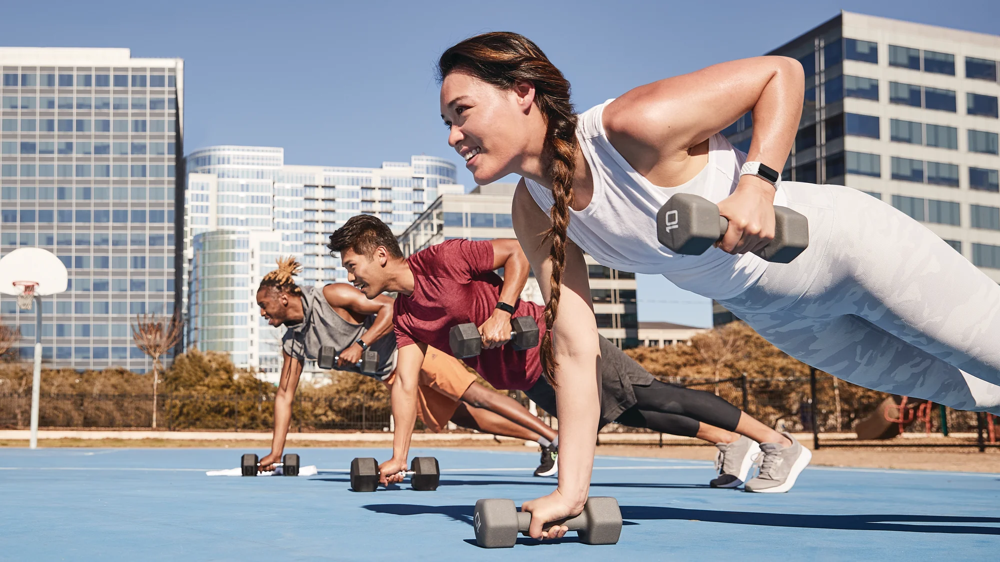 Three young people in a line on a basketball court, wearing athletic clothes and doing plank rows with weights, while wearing Fitbit Charge 5 trackers.