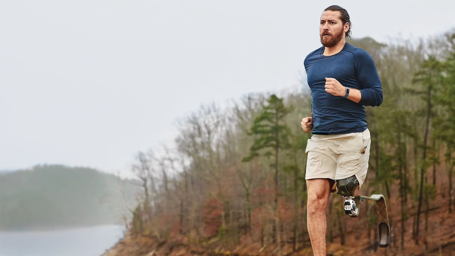 A young man in a blue shirt goes for an outdoor run next to trees and a body of water while wearing a Fitbit Charge 5.