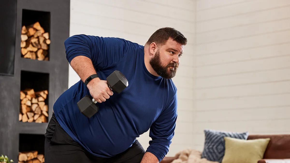 A man in a blue shirt and black pants doing one-arm dumbbell rows while wearing a Fitbit Charge 5.