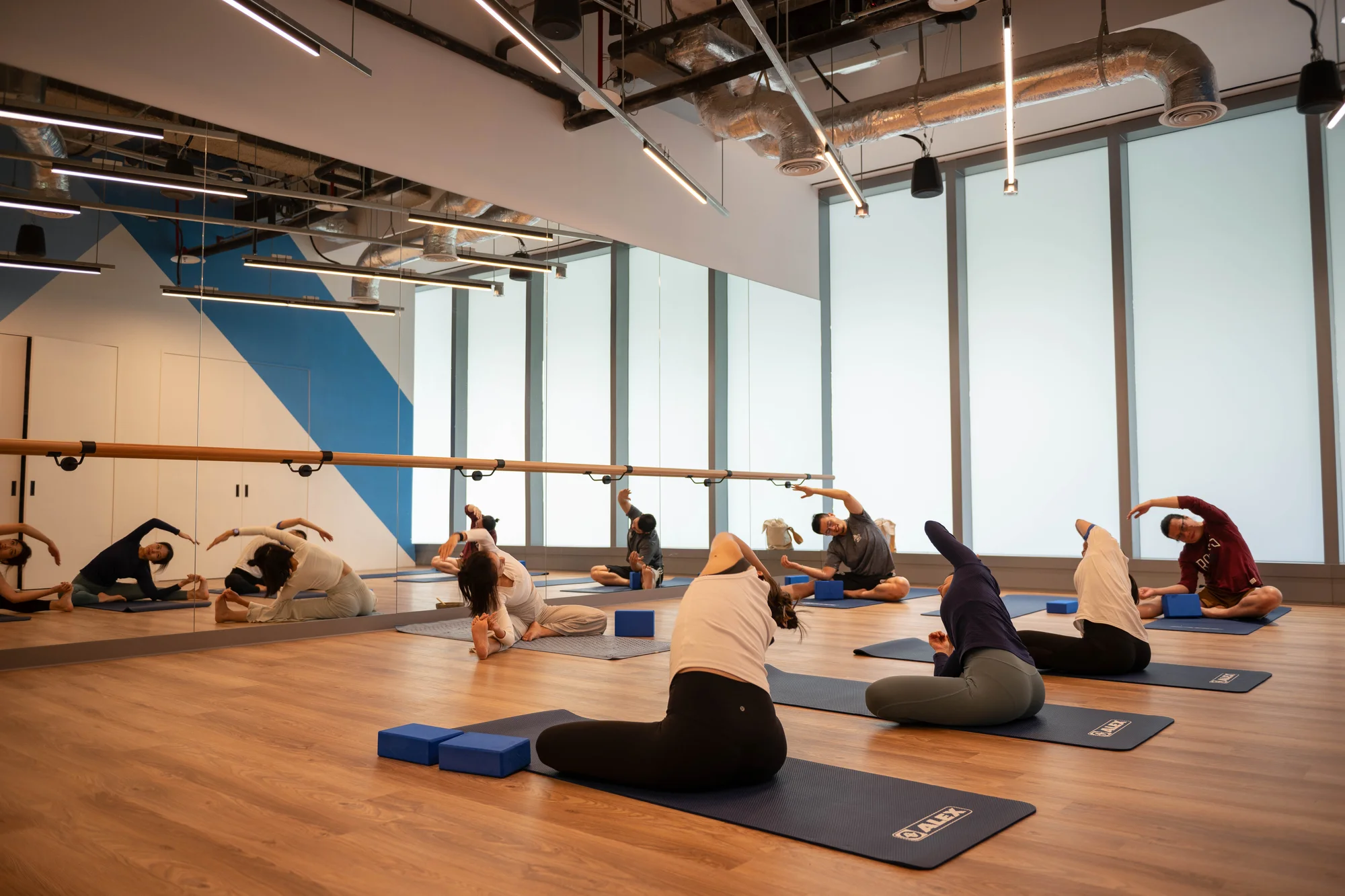 A group of people on yoga mats sit cross legged and stretch in a room with wooden floors and a mirrored wall.