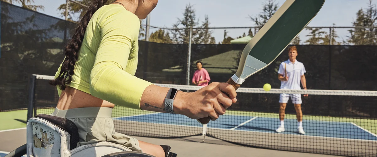 A photo of three people playing pickleball. There are two people in the background on one side of the next. The person in the foreground is in a wheelchair and is wearing a Fitbit device on their wrist.