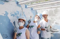 Image of three volunteers standing in front of a wall, wearing helmets and a Just A Change t-shirt and holding putty knives.