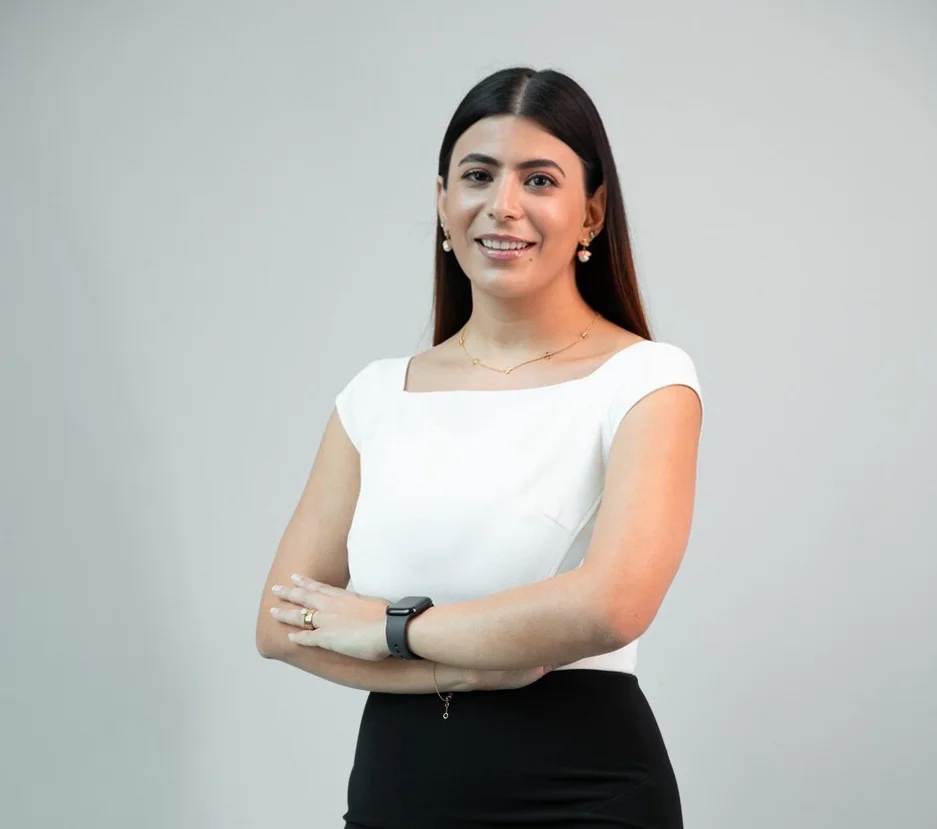 A woman wearing a white shirt and black pants stands in front of a grey background, with her arms folded.