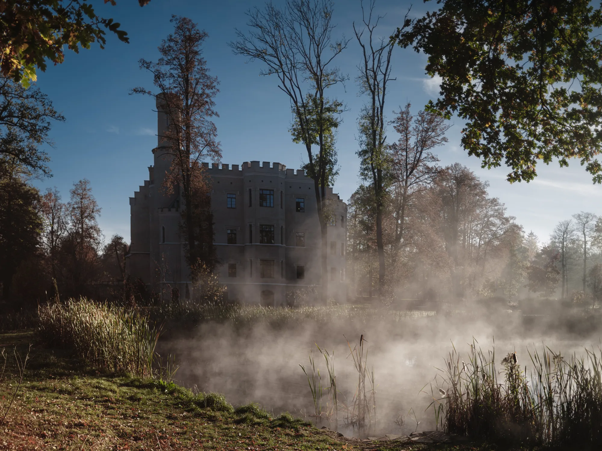 In the photograph, we can see a small pond and a morning fog. In the center, there is a big castle, covered in this fog. The sky in the background is blue. There are a few trees in front of the castle. It looks as if the day is starting.