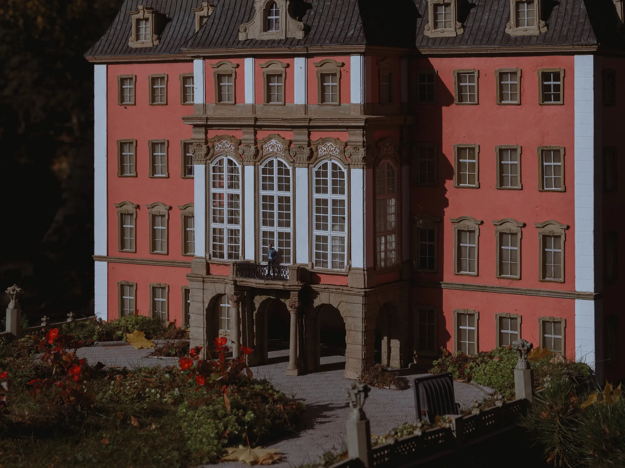 The whole space of the photograph presents the front wall of a big, old castle. The facade is in two colours: light brick-like and white. There are six rows of windows, varying in shape. The three biggest windows are located at the center of the castle front wall. The castle casts a long, dark shadow, suggesting it’s evening time.