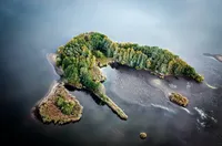 Ponds seen from a bird's eye view. Dark blue and navy blue water. In the center of the photo you can see an irregularly shaped island, all covered with trees. The trees are in various shades of green. The lightest trees have a slightly yellowish hue. Light reflections of the island are visible in the waters.