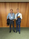 Two men, Yossi and Anthony, stand next to each other against a wood-paneled wall.