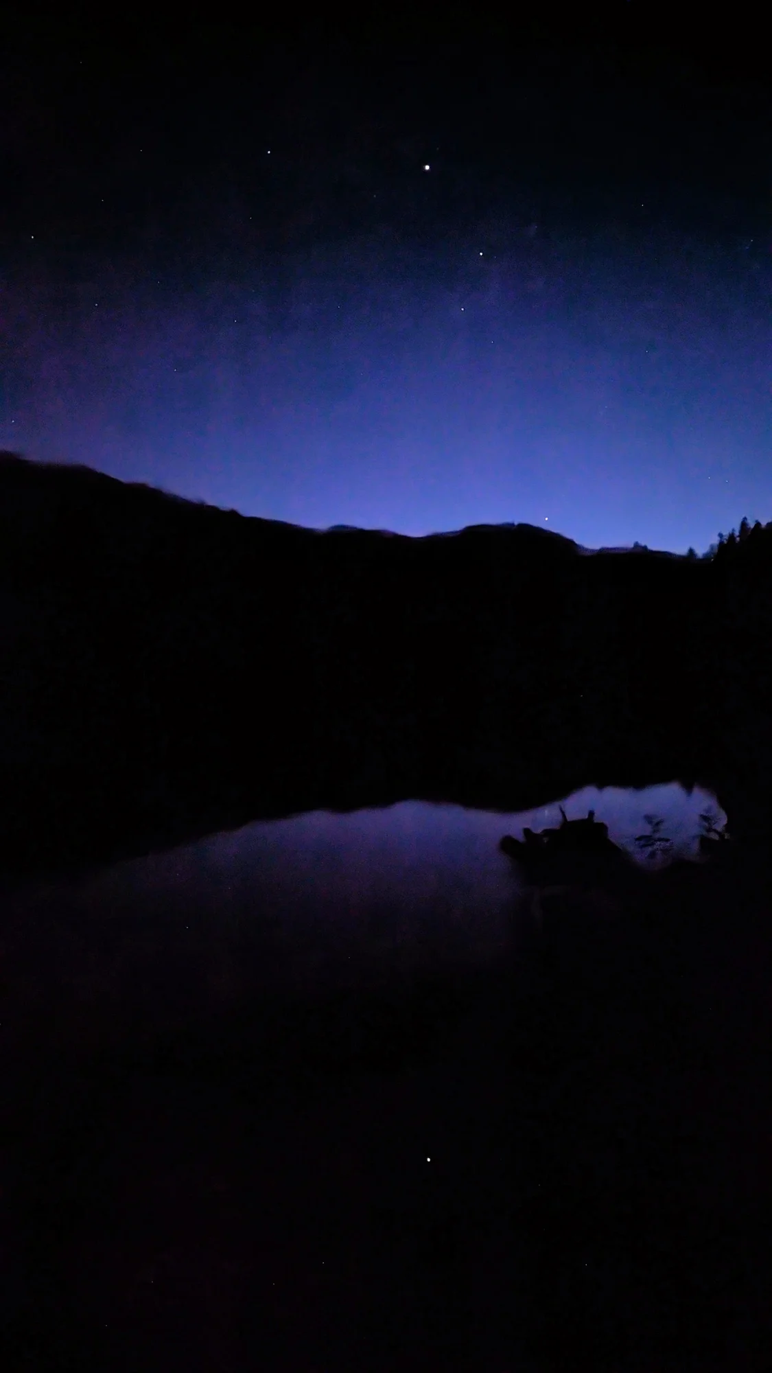 A photo of the night sky. There are stars above a dark blue lake and in the background, the silhouette of a mountain range.