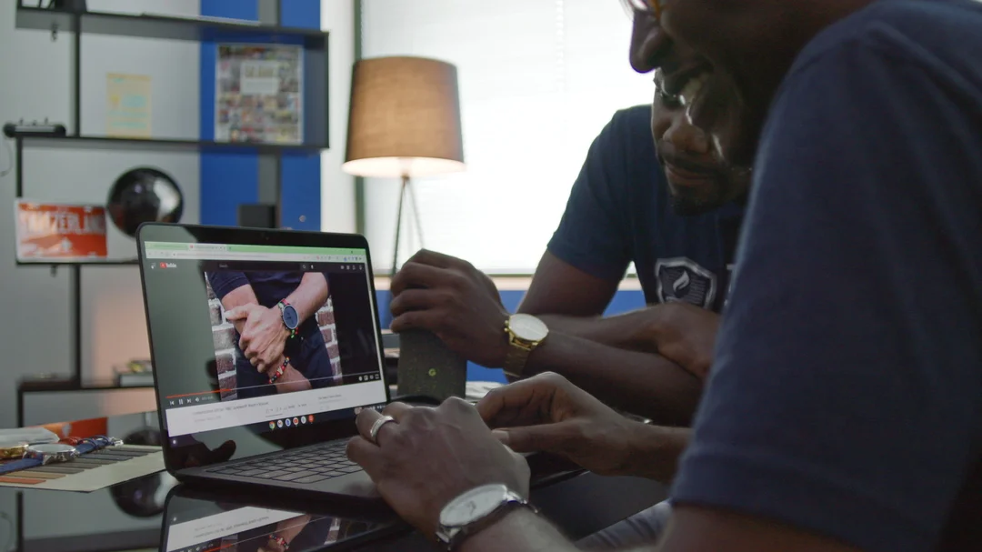 Two men sit at desk looking at a laptop computer showing a YouTube video that shows a person’s hand wearing a watch