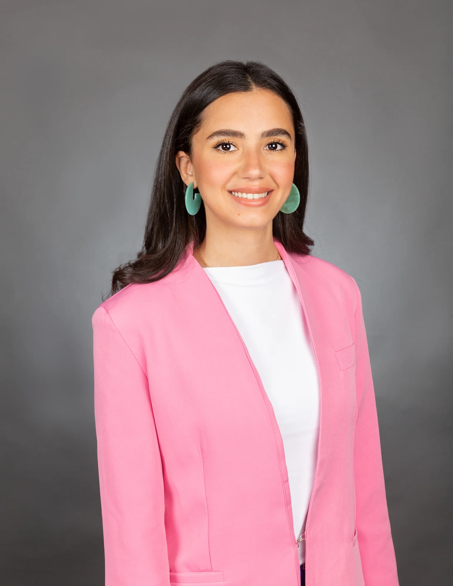 A woman with long black hair and wearing a white shirt, pink blazer and turquoise hoop earrings smiles at the camera.