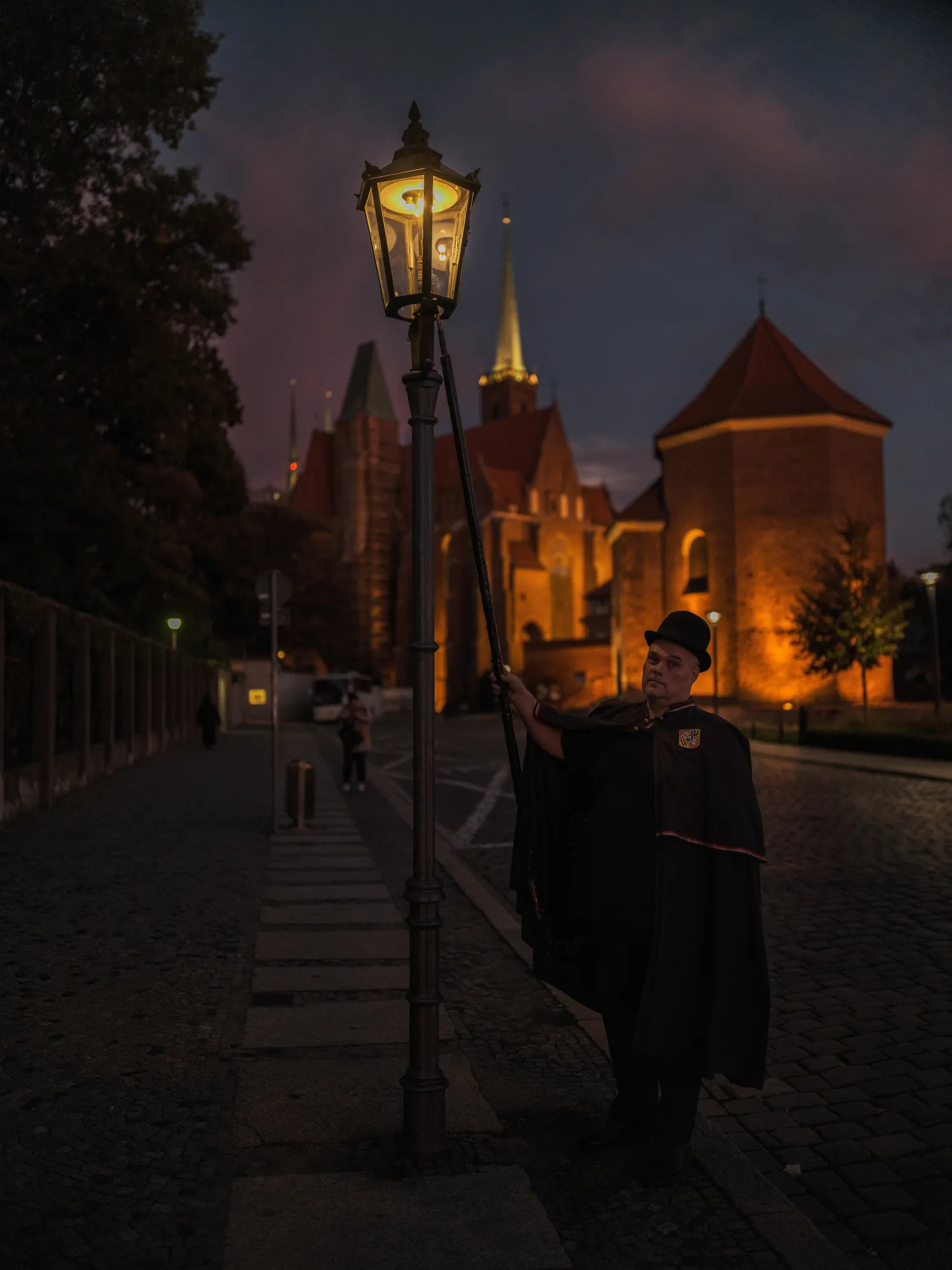 The photo depicts a nighttime scene featuring a man dressed in a traditional outfit, lighting a gas lamp on a cobblestone street. The man is wearing a dark cloak and a hat, and he is using a long pole to ignite the lamp. The background includes historic buildings illuminated by warm lighting, which contrasts with the darkening sky. The street is lined with trees and other lampposts, creating a picturesque and old-fashioned atmosphere.