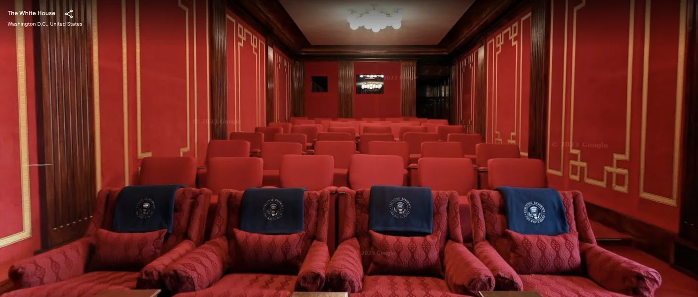 A street view capture of the movie theater located in the White House. The walls of the room are adorned by red and gold panels. There are several rows of seats, all of which are red. The four in the front row are still different, but distinct from the rest through their quilted red upholstering. Each one has a navy blue covering over the head of the seat.