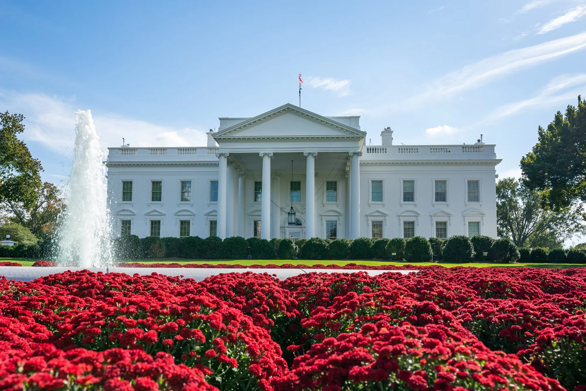 A color photograph of the White House’s front view. It is day time and the sky is blue. The water flows out of the fountain in the front, in this image, found on the left. Green bushes surround the front lawn of the White House, and to the side, there are green trees. Across from the bushes are a large garden of red flowers.