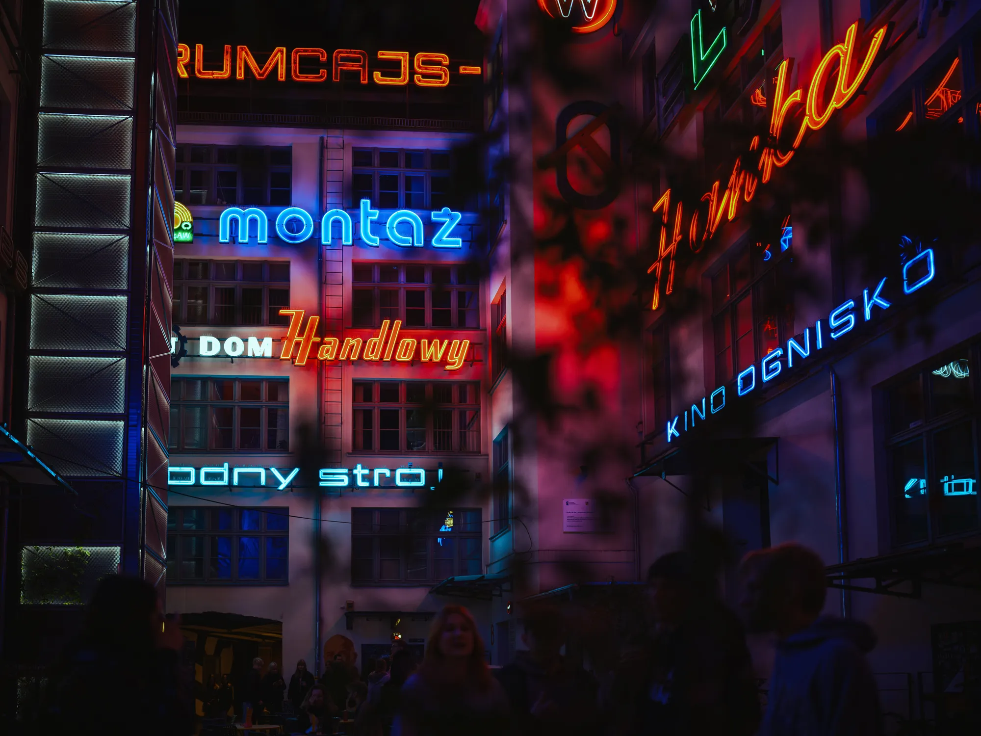 The photo depicts a vibrant night scene featuring an array of colorful neon signs in various styles and colors. The signs are attached to the facades of buildings, which appear to be part of a street or alleyway. The signs display text in what seems to be Polish, including words like "montaż," "Handlowy," and "Kino Ognisko." The lighting from the neon signs creates a striking and colorful glow, casting reflections and shadows on the surroundings.