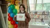 A young artist with short brown hair and a brown sweater is holding a certificate with her artwork of family and friends enjoying dinner. Behind her is a larger version of the artwork alongside a cluster of yellow, red, blue and green balloons.