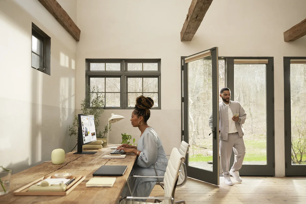 A woman sits and types on a computer at a wooden desk with a new Nest Wifi Pro displayed. Behind her, a man opens a glass door while looking at his phone.