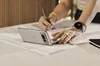 A Porcelain Pixel Fold sits in a half-opened, clamshell position on a desk on top of papers. A person’s hands are nearby, one lightly tapping one half of the opened screen of the Fold and the other holding a pencil and sketching on the papers underneath it.