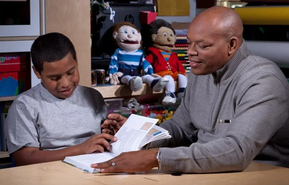 Picture of a man sitting beside a young child, assisting them with their studies. Both the man and the child look at an open textbook.