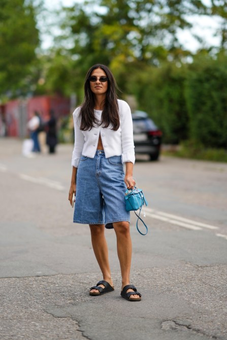 Womam wears black sunglasses, a white tweed buttoned jacket, blue denim large jeans shorts, a blue shiny leather Puzzle handbag from Loewe, black matte leather belted sandals from Birkenstock, outside Rabens Saloner, during Copenhagen Fashion Week